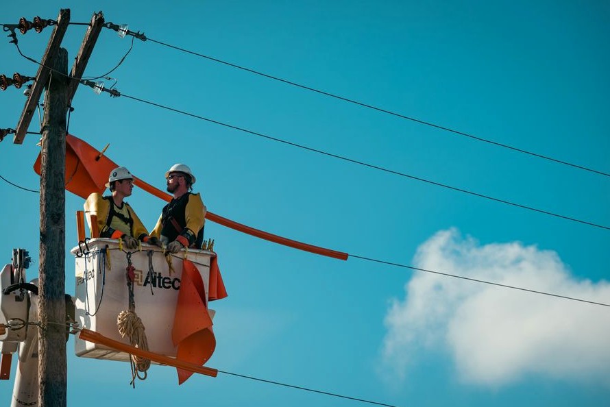 Two electricians in safety gear working on a utility pole with a clear blue sky backdrop